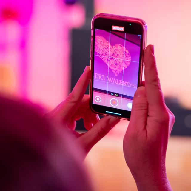 Zbliżenie: osoba trzyma telefon i nagrywa scenę z napisem „Koncert Walentynkowy”.